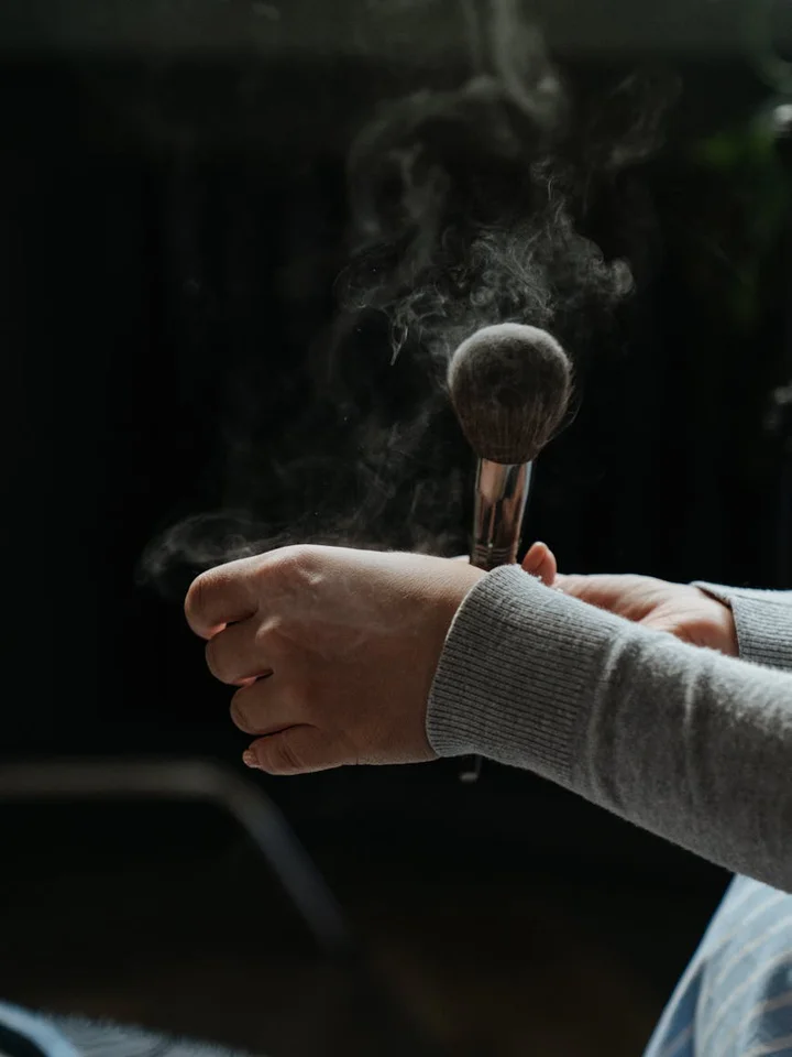 A hand holds a makeup brush with mist or steam in the background, prepared for makeup application.