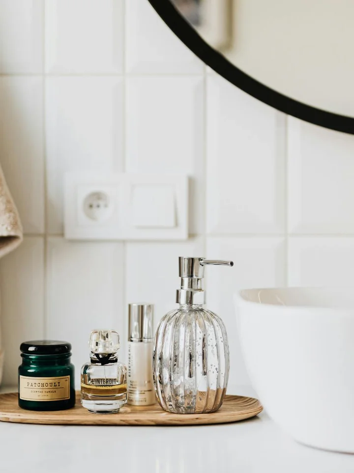 A bathroom vanity with a wooden tray holding small skincare bottles and a glass pump dispenser, beside a white sink and a round mirror.