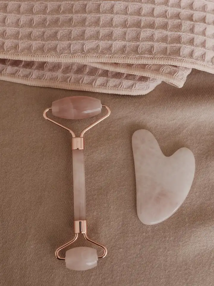 Rose-quartz facial roller and a gua sha stone on a soft beige towel, representing common at-home tools for applying serums and optimizing skincare routines.
