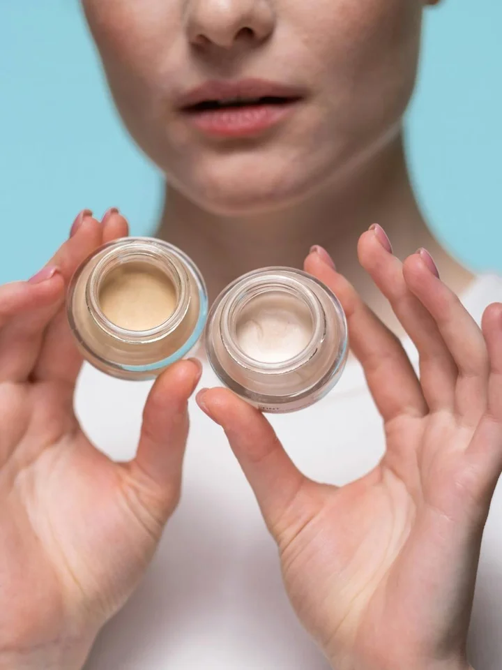 Close-up of a person holding two small jars of skincare cream against a light blue background.