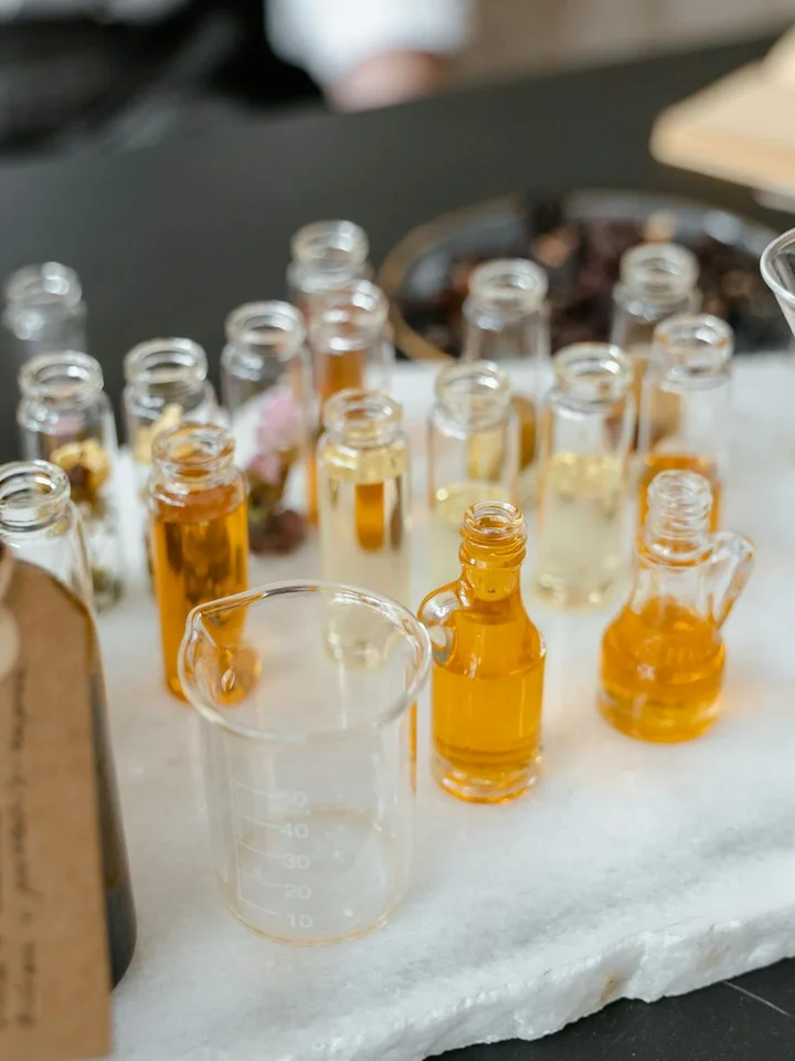 Assorted small amber glass bottles with golden oils arranged on a white marble surface, with a clear beaker in the foreground, suggesting essential oils for skincare.