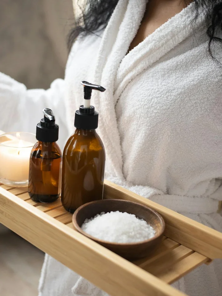 Spa-style setup with amber castor oil bottles on a wooden tray, a bowl of salt, and a person wearing a white bathrobe ready for facial care.