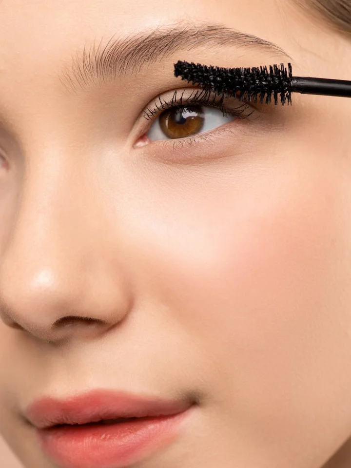 Close-up of a woman's eye with a mascara wand hovering over the upper lashes.