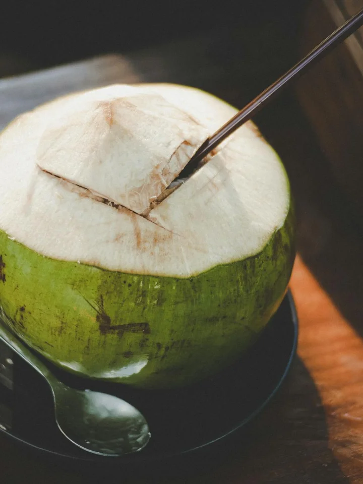 Fresh green coconut with the top cut open, a metal tool inserted, resting on a wooden surface.