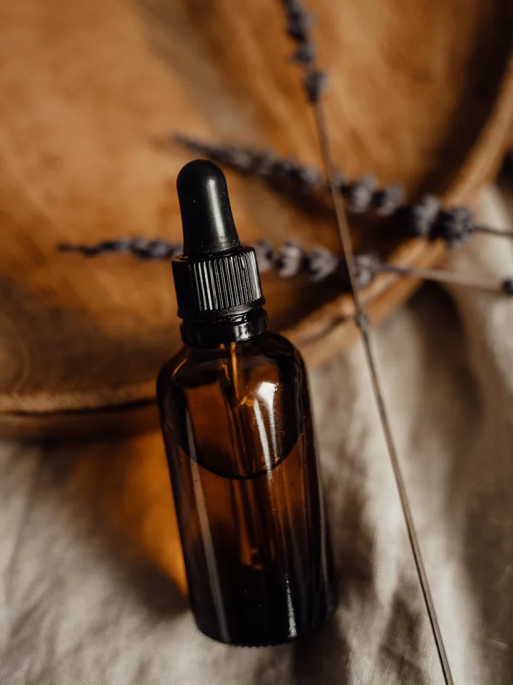 A brown glass dropper bottle with a black cap sits on a soft fabric, with dried botanical stems in the blurred background.
