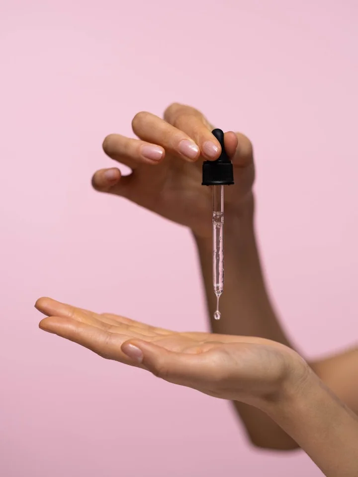 Hands hold a dropper releasing a clear serum drop onto an open palm against a pink background.