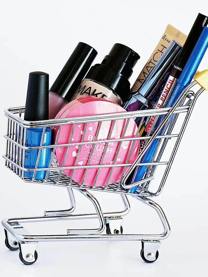 Close-up of a metal shopping cart filled with makeup bottles, tubes, and brushes, illustrating evaluating product claims and INCI lists.