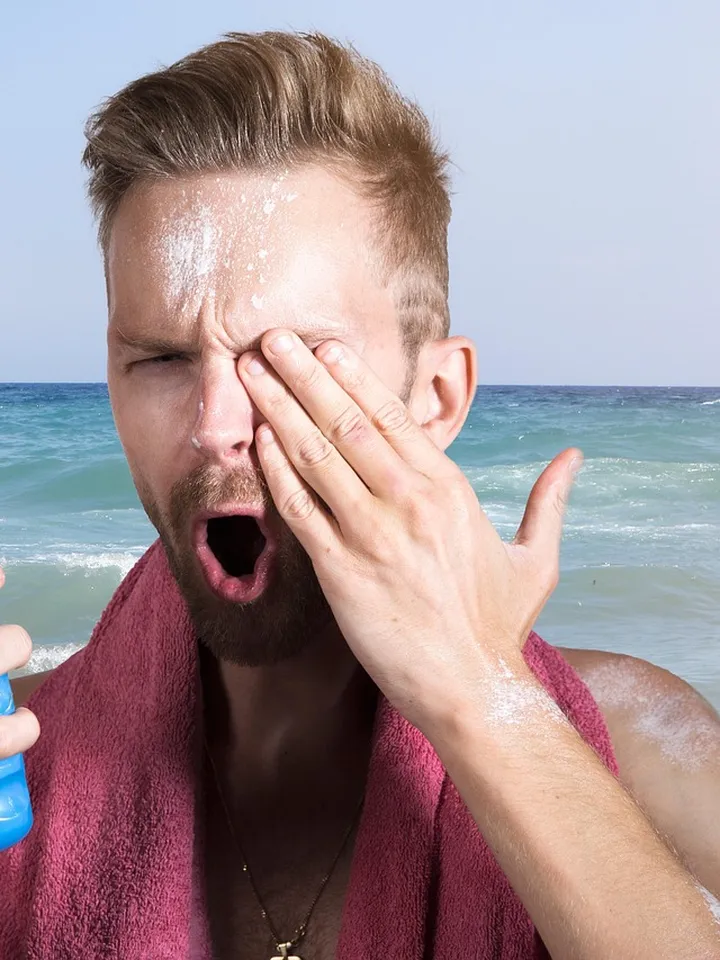 Man at the beach applying sunscreen spray and touching his face.