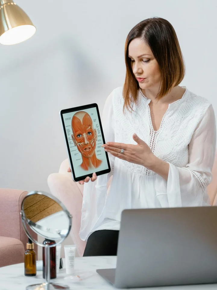 A woman sits with a laptop, holding a tablet that displays a facial diagram during a skincare consultation.