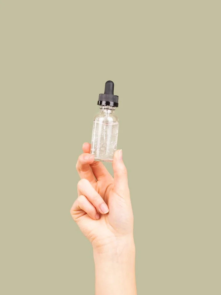 A hand holding a small glass dropper bottle with a black cap against a beige background.