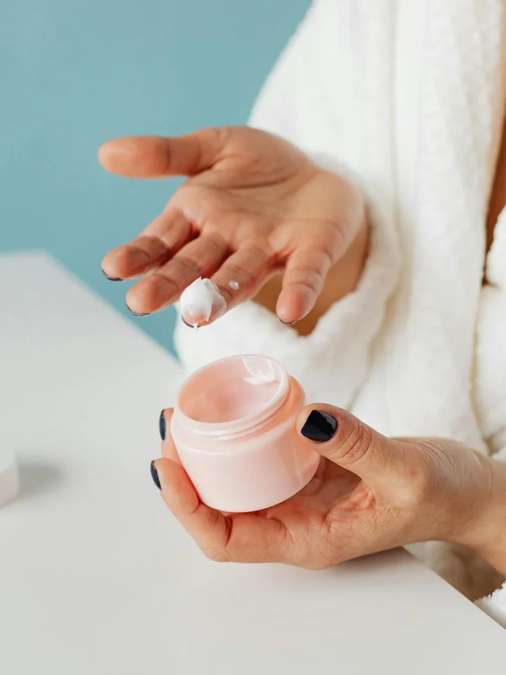 Close-up of hands applying a small amount of moisturizer from a pink jar onto fingertips, with a soft white robe and a light blue background.