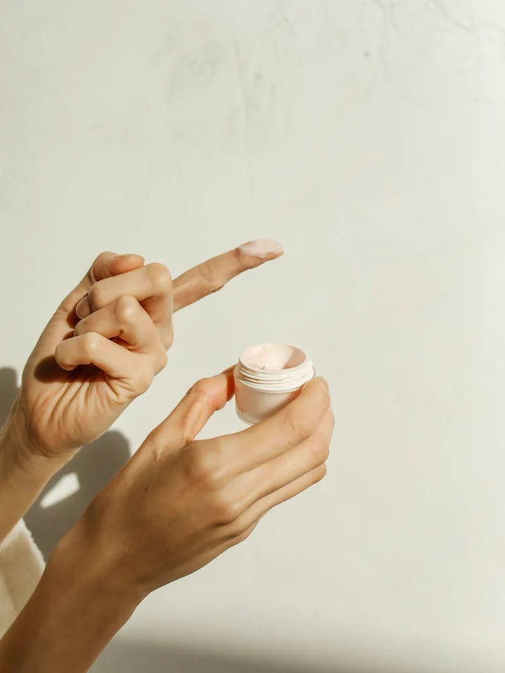 Close-up of hands holding a small jar of cream, with one finger dipped to scoop out moisturizer.