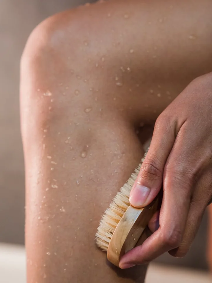Close-up of a hand holding a natural-bristle brush and applying it to damp skin on the arm, demonstrating physical exfoliation.