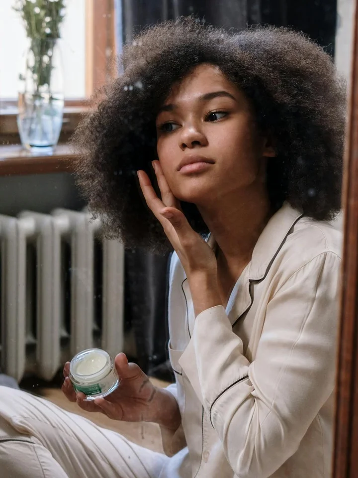 Person with curly hair wearing a light-colored jacket sits indoors, holding a small jar of skincare product and preparing to apply it.