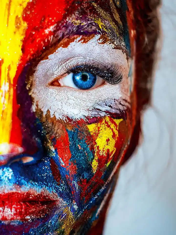 Close-up of a person's face covered in multicolored paint with a bright blue eye visible.