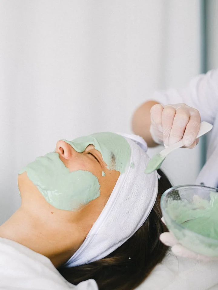 Close-up of a person lying back while a green facial mask is applied by a practitioner.
