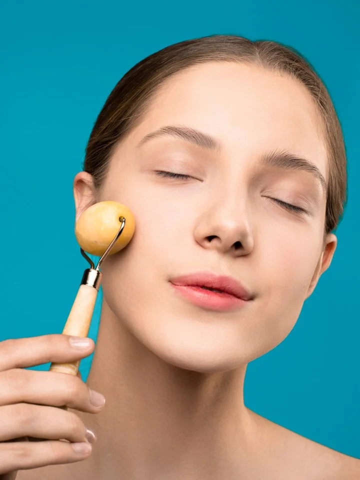 Close-up of a person with eyes closed using a facial roller on the cheek against a blue background