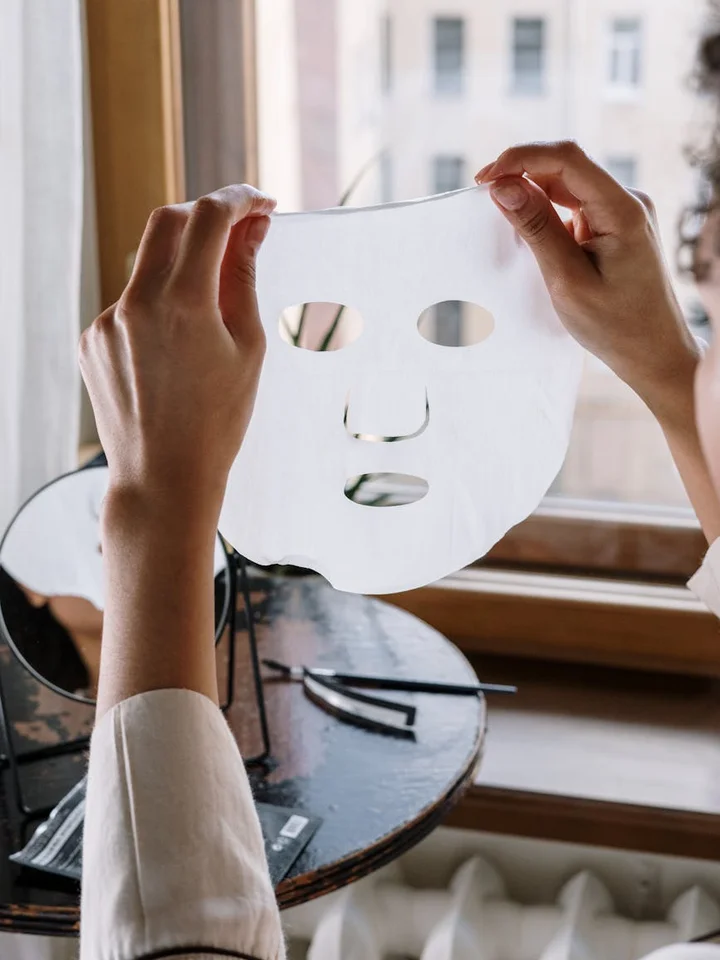 Hands holding a white sheet facial mask near a window over a vanity mirror, about to apply it.