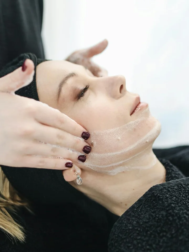 Close-up of a skincare professional applying a sheer mask or gel on a woman's face during a facial treatment; the client lies back with eyes closed.