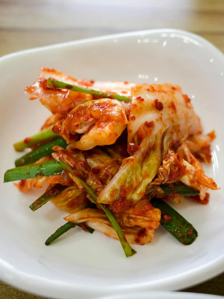 Close-up of kimchi in a white bowl, showing fermented cabbage and greens with red pepper seasoning.