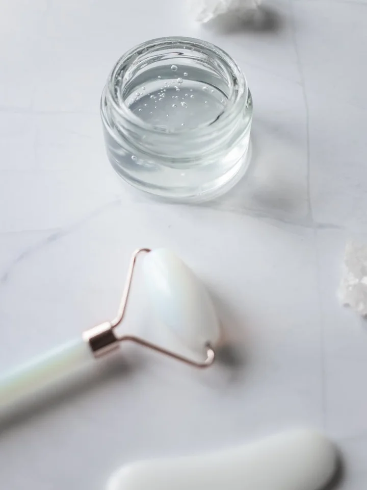 Close-up of a glass jar with clear serum on a marble surface and a small curved skincare tool in the foreground