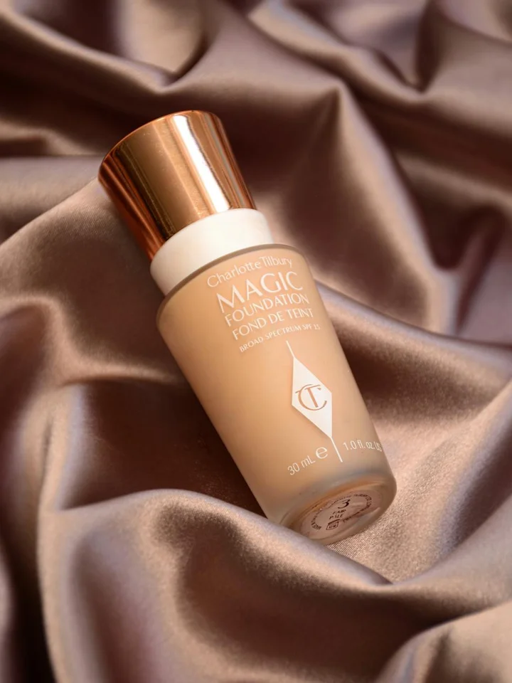 Close-up of a beige liquid foundation bottle with a copper cap resting on soft brown satin fabric.