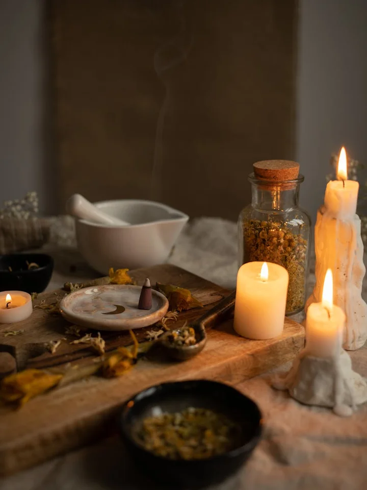 Moody still-life of a wooden board with dried botanicals, a mortar and pestle, glass jars, and lit candles, illustrating preparation and extraction themes.