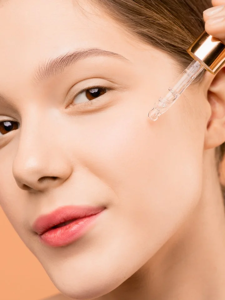 Close-up of a woman with clear skin holding a glass dropper near her cheek, preparing to apply skincare serum.