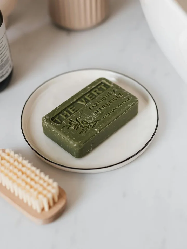 Green rectangular soap bar on a small white plate with a gold rim, placed on a marble surface, with a beige toothbrush in the foreground.