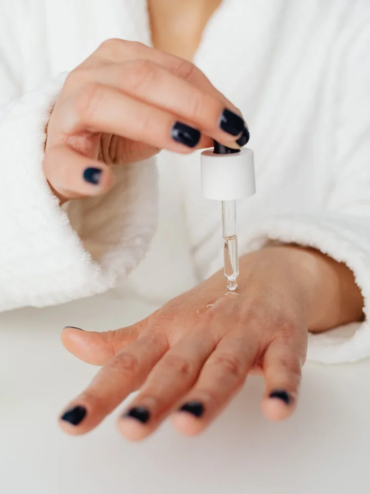 Close-up of a hand with a dropper applying skincare serum, wearing a white robe and dark nail polish.