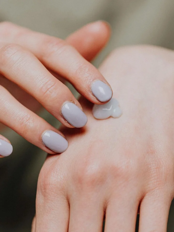 Close-up of a hand with lavender nail polish applying a dollop of white moisturizer to the back of the hand.