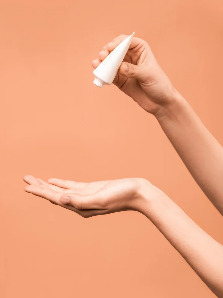 One hand holds a small white squeeze tube above the other open palm, as if dispensing cream, against a peach-colored background.