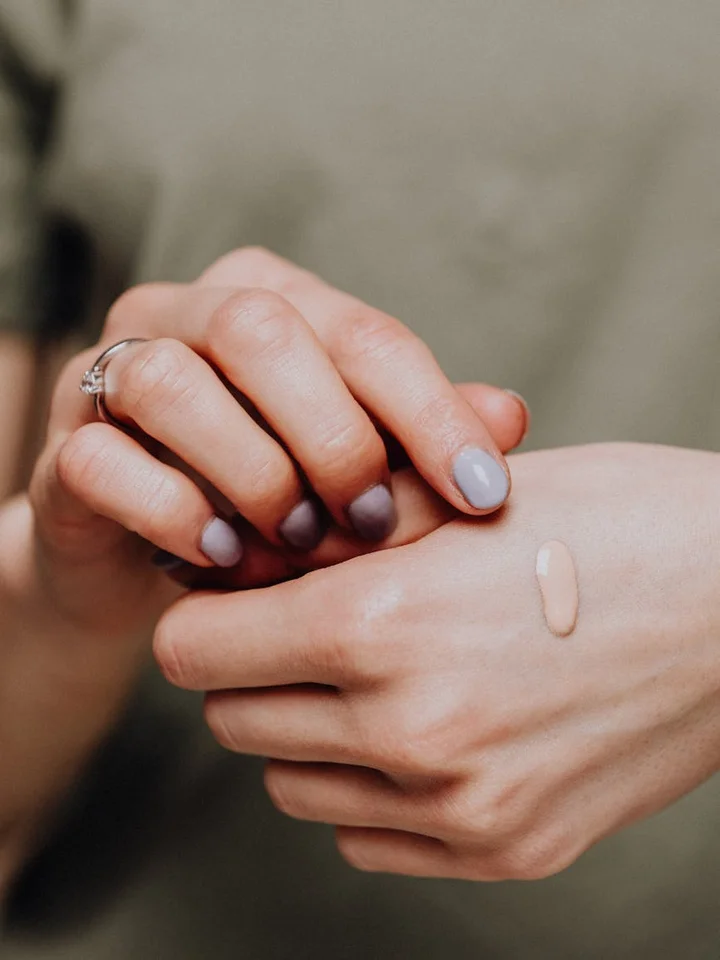 Close-up of hands with a small foundation swatch on the back of one hand; nails painted in soft gray.