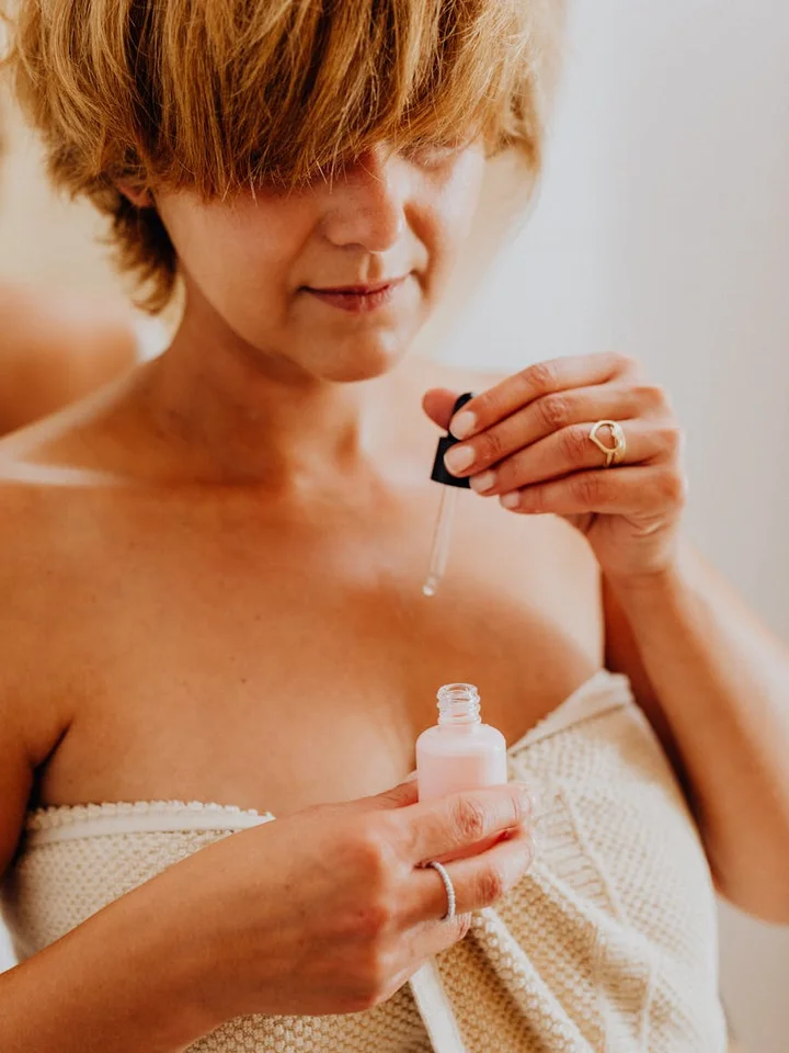 A person wearing a towel applies hyaluronic acid serum from a dropper bottle, focusing on the neck/upper chest area.