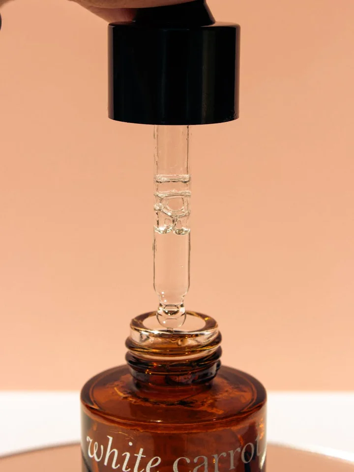 A dropper hangs over a brown glass bottle of serum against a warm peach background.