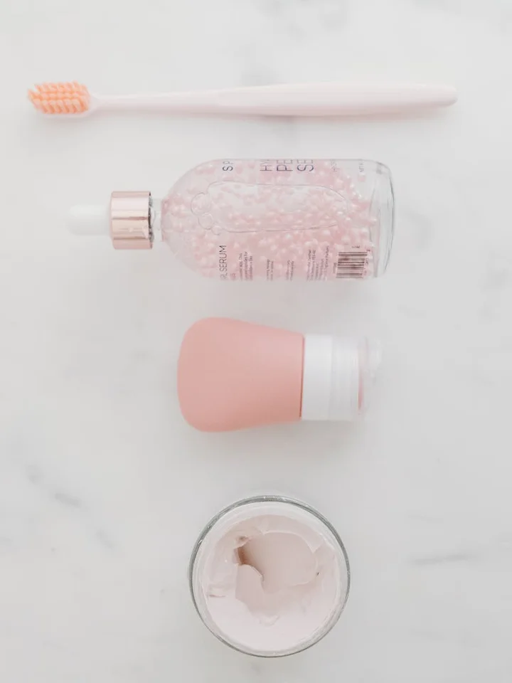 Flat lay of skincare items on a white surface: a pink toothbrush at the top, a glass dropper bottle with pink liquid, a pink makeup sponge, and an open jar of pale pink cream.