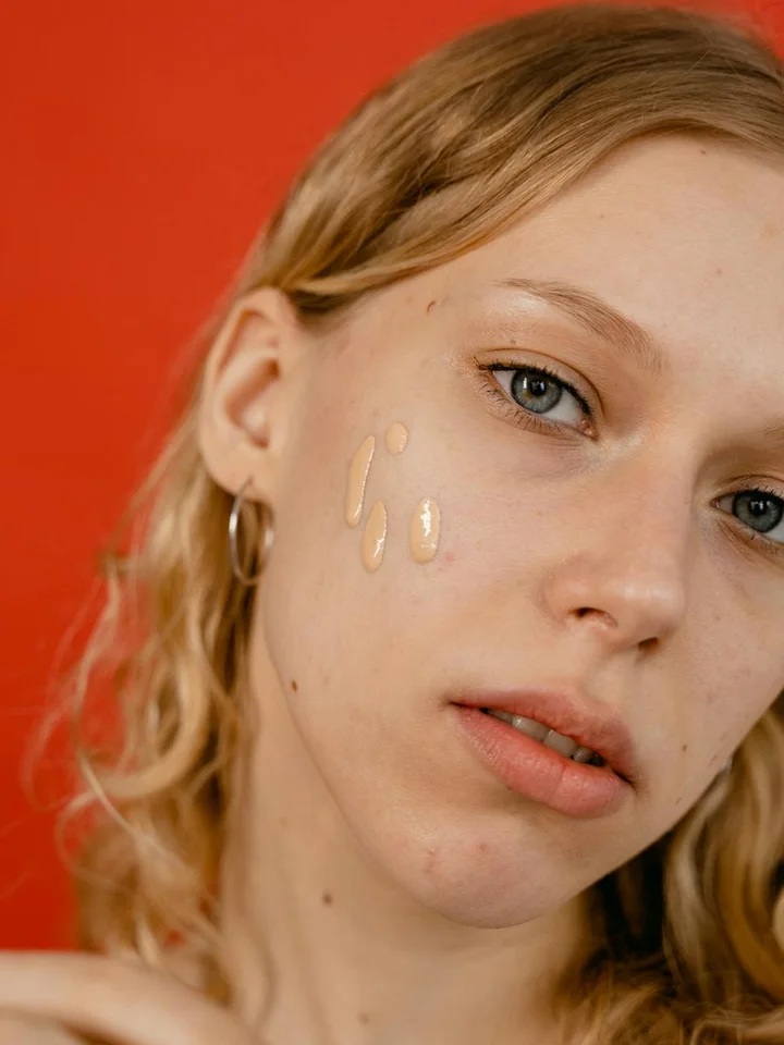Close-up of a young woman with foundation swatches on her cheek against an orange background.