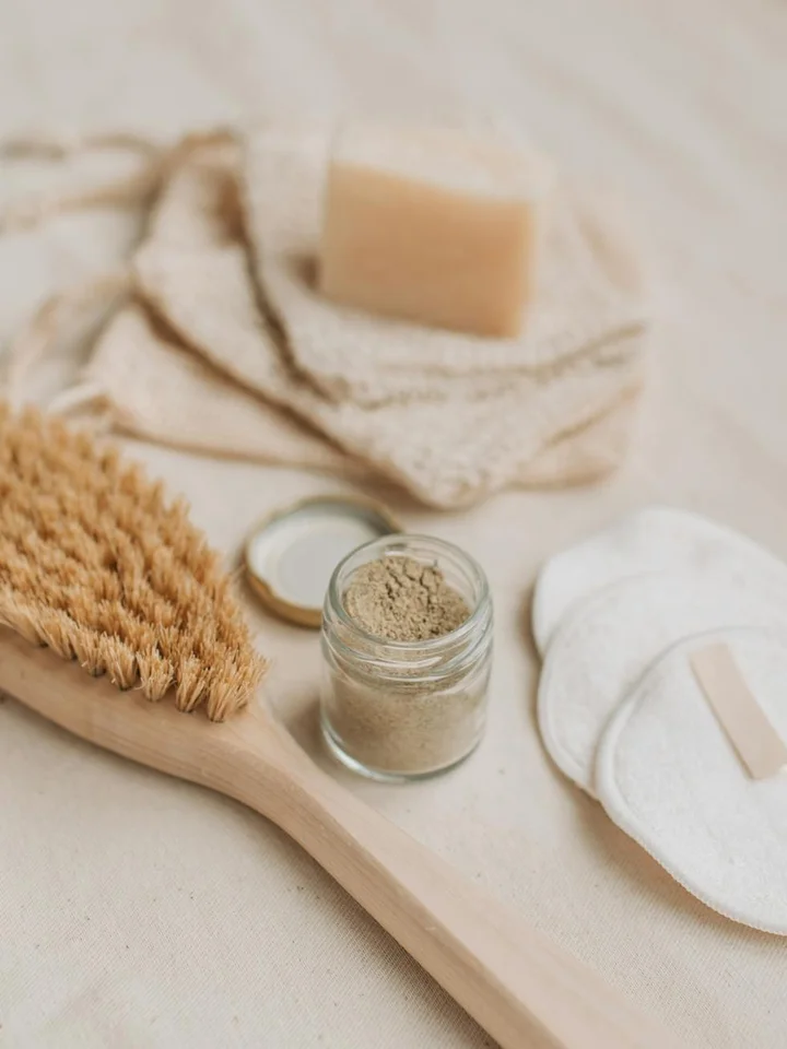 Natural skincare setup featuring a wooden body brush, a small jar of powder, cotton pads, and a bar of soap on a light beige surface