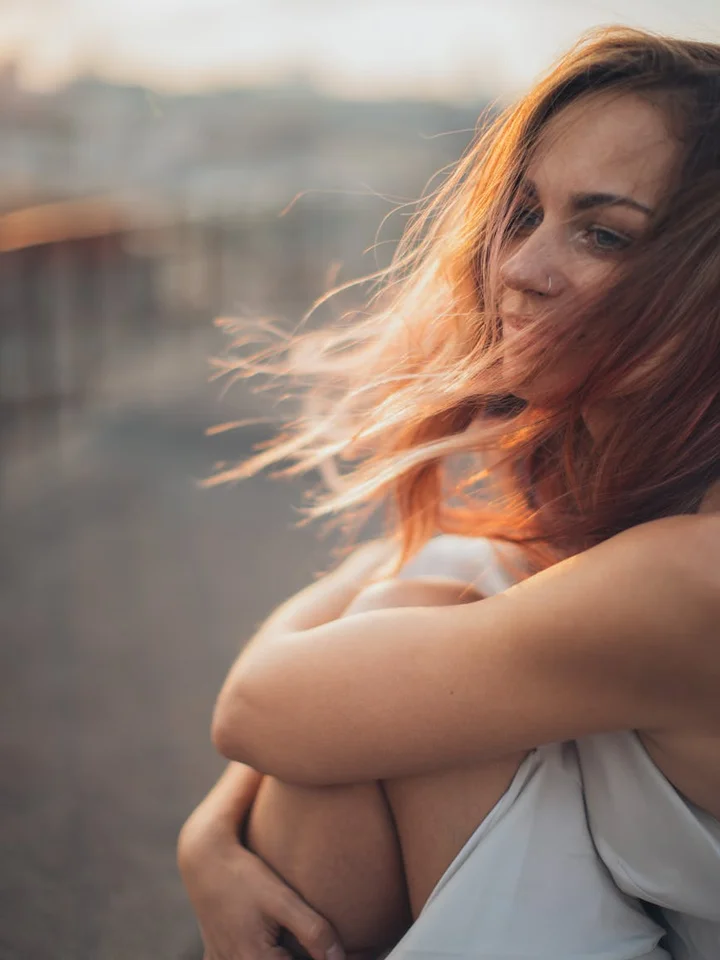 Portrait of a woman with wind-blown hair sitting outdoors, arms wrapped around her knees, looking toward the camera.