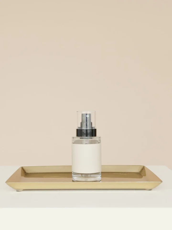 A clear serum bottle with a pump sits on a gold tray against a pale beige background.