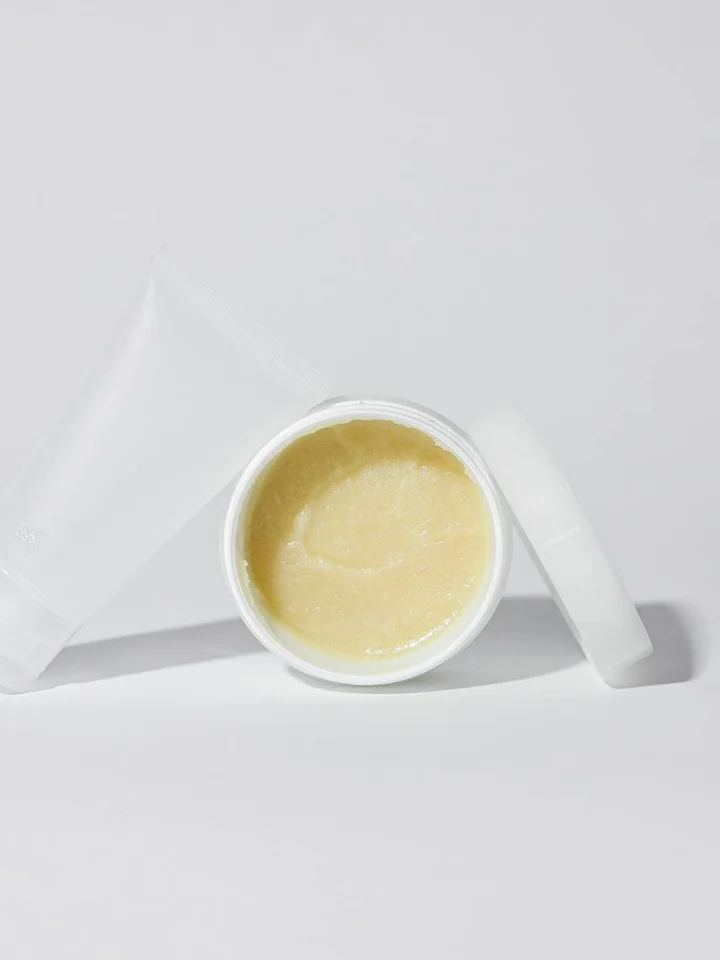 Open jar of pale yellow cream with a white lid placed to the side on a white background.