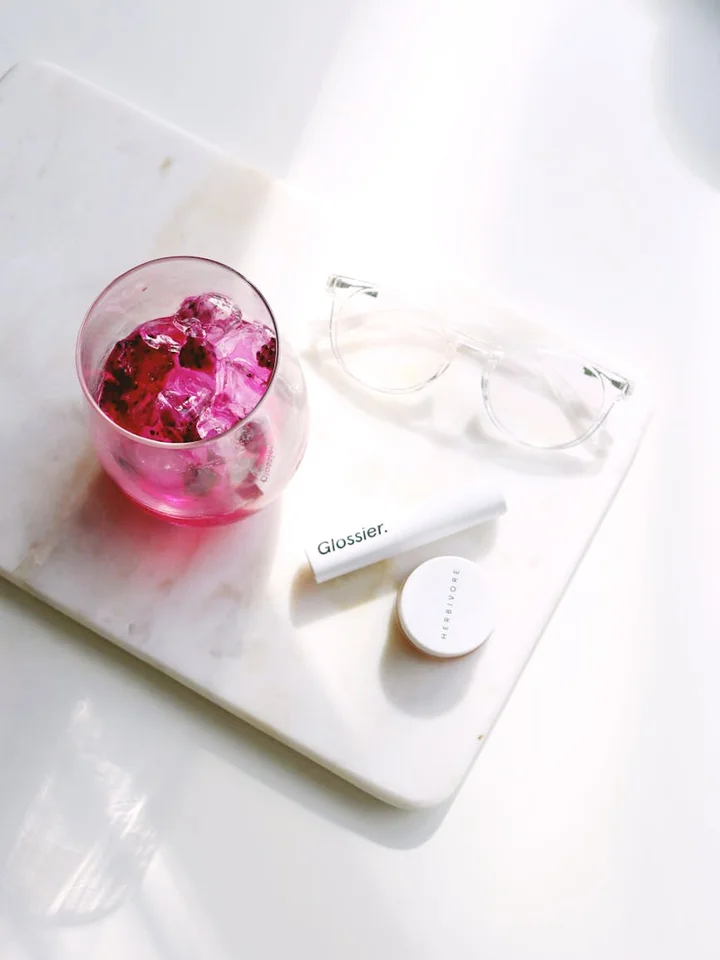 A flat lay of beauty items on a white marble tray: a pink glass with pink crystals, a white Glossier tube, a small white container, and a pair of clear-framed glasses.
