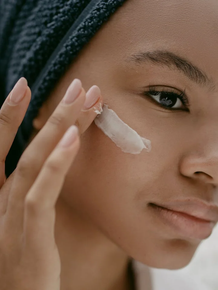 Close-up of a person with a towel wrapped around their head applying a cream to their cheek, illustrating skincare routine.