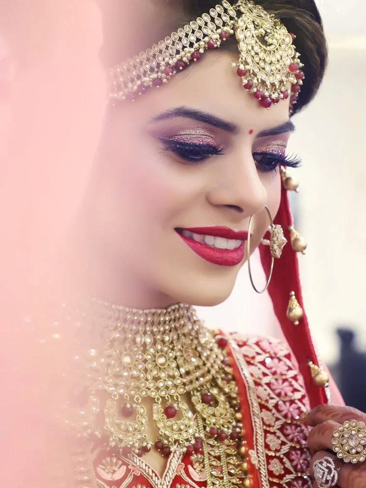Close-up portrait of an Indian bride wearing ornate gold jewelry and bold makeup, including red lipstick and shimmering eyeshadow