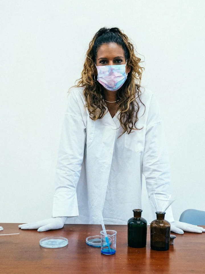 Cosmetic chemist wearing a white lab coat and a face mask standing at a lab bench with bottles and beakers