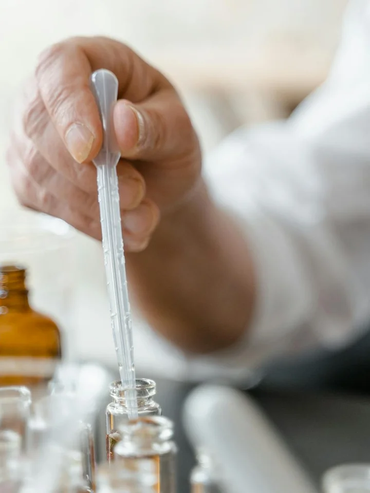 Close-up of a hand using a glass dropper to transfer liquid into small glass vials in a laboratory setting.