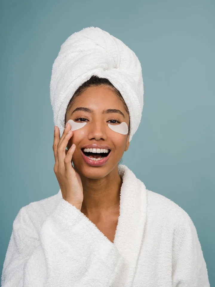 Smiling woman with a towel wrapped around her head and under-eye patches during a skincare routine.