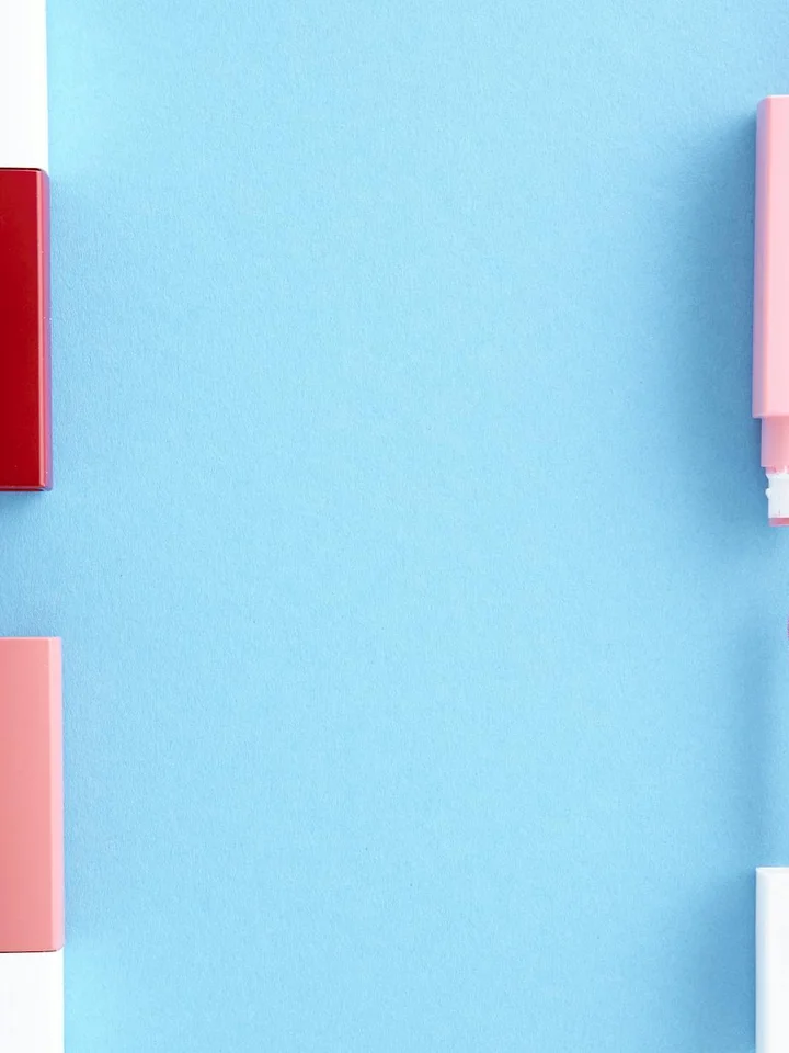 Flat lay of several lipstick tubes with red, pink, and white caps placed along the edges of a bright blue background.