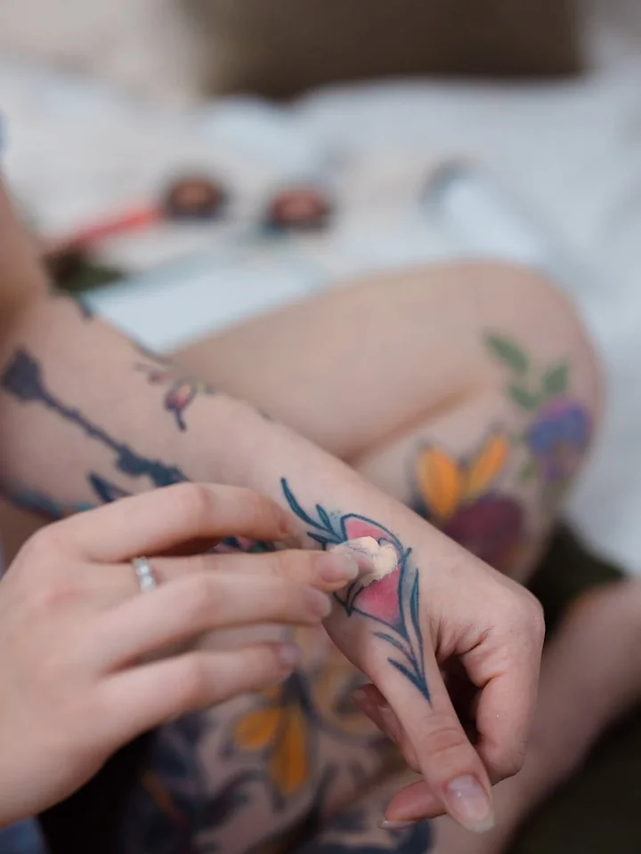 Close-up of tattooed hands with a ring, handling makeup in a soft-focus setting.