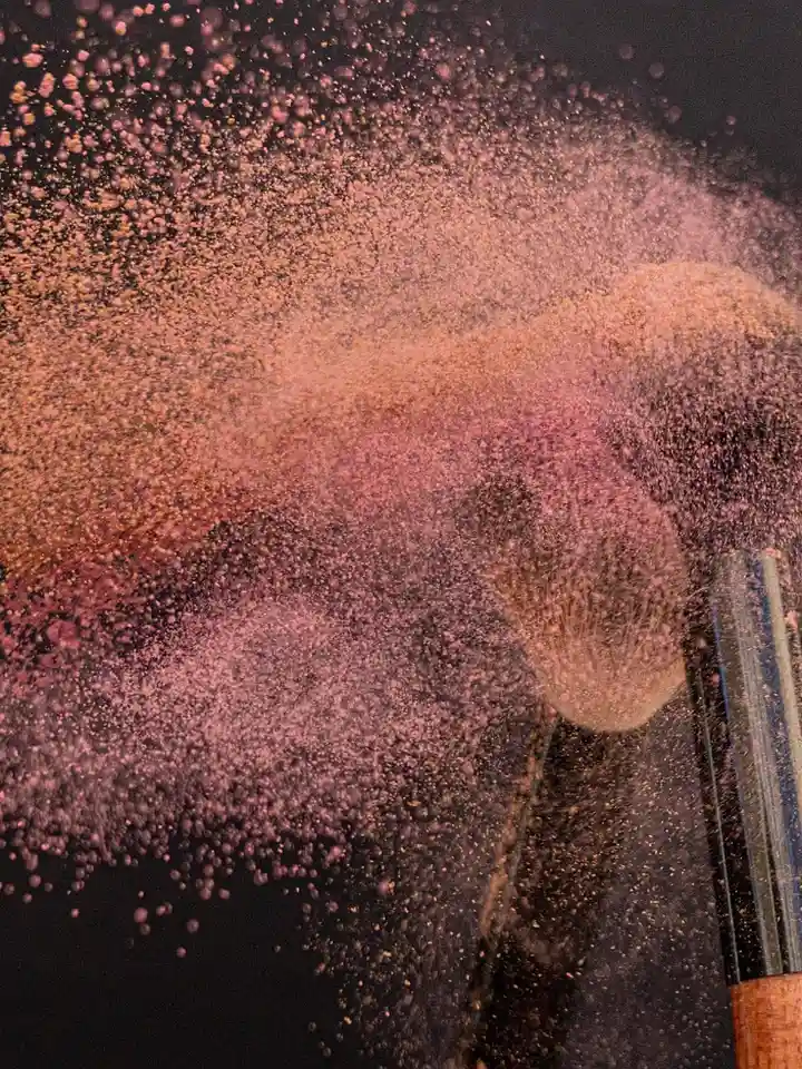 Close-up of pink cosmetic powder dispersing into the air against a dark background, reminiscent of MAC foundation textures.